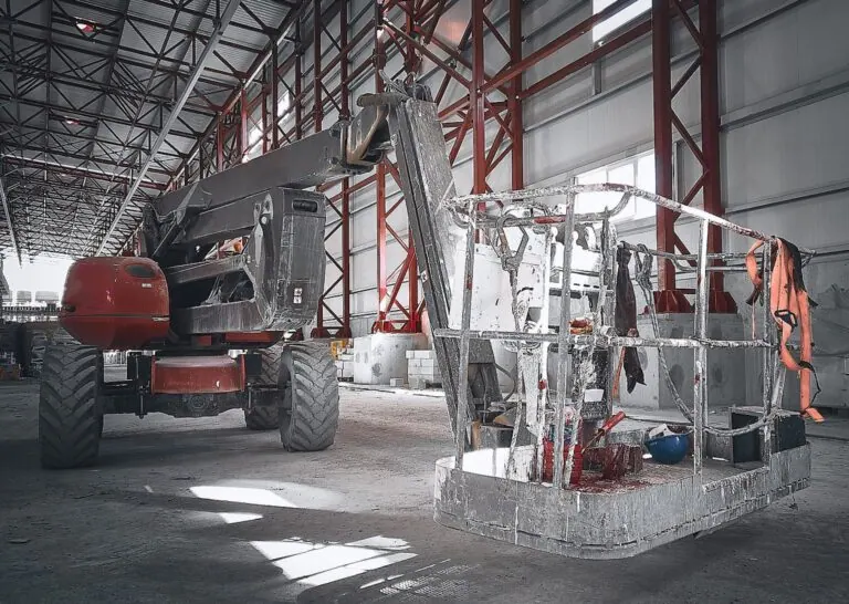 Aerial lift platform with a safety harness hanging inside a large industrial warehouse with red steel beams and a high ceiling.