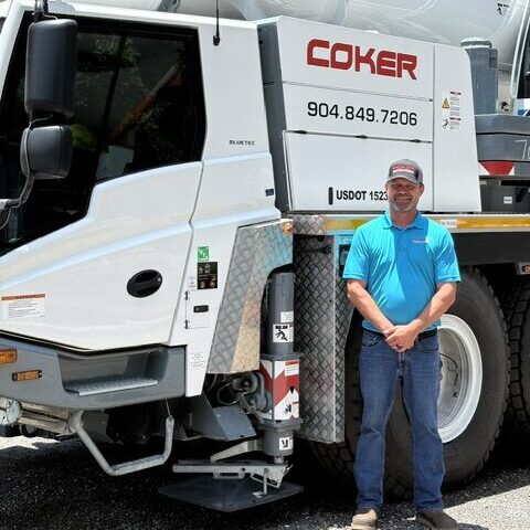 Man in blue shirt and jeans standing next to a large white Coker crane truck with a person in the cab.